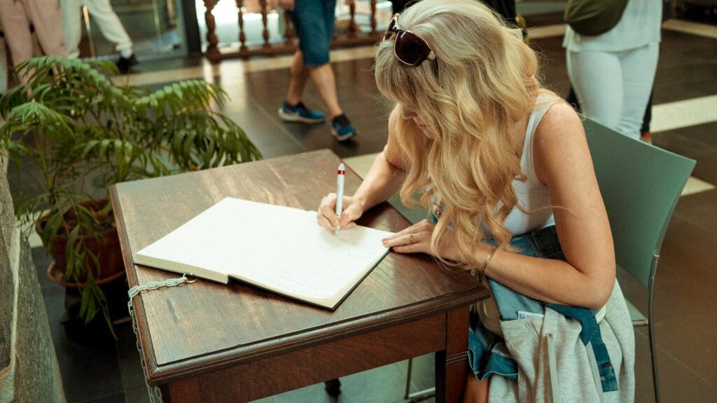 A woman with long blonde hair writes in a journal at a table indoors in Ghent, Belgium.