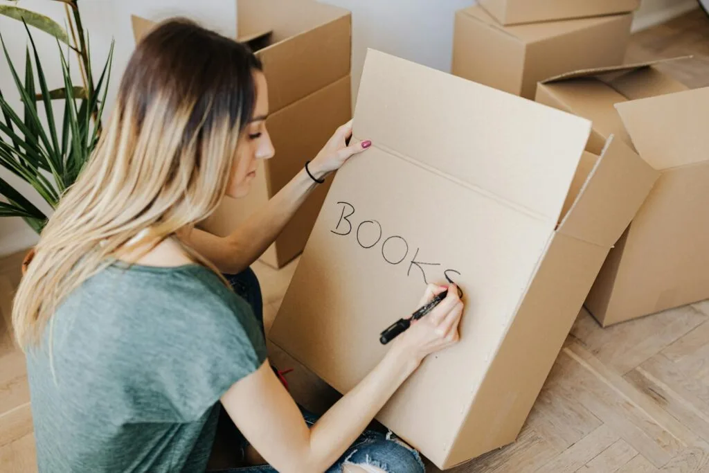 Woman writing on cardboard box while packing books, preparing for moving day at home.